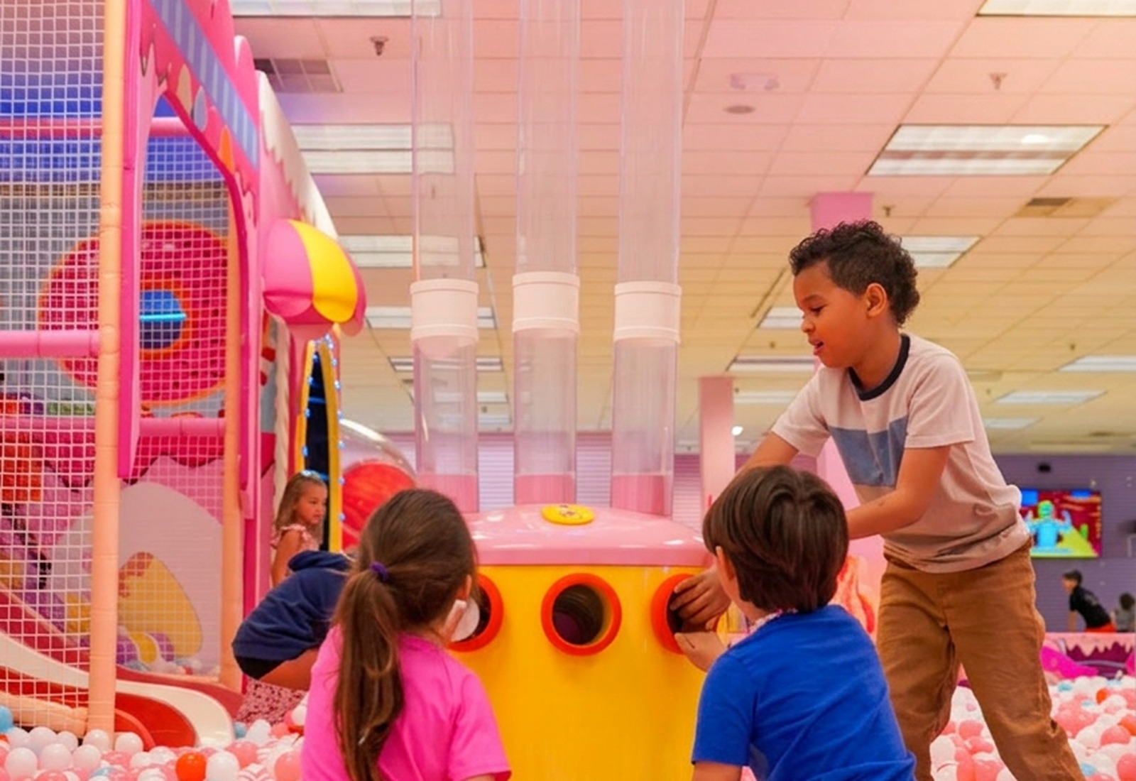 Three young kids playing in a giant pastel pink, white and blue ball pit around a yellow ball cannon under a glowing UFO-shaped ball dropper at Star Park indoor playground